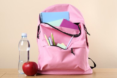 Photo of Backpack with different school stationery, bottle of water and apple on wooden table against beige background