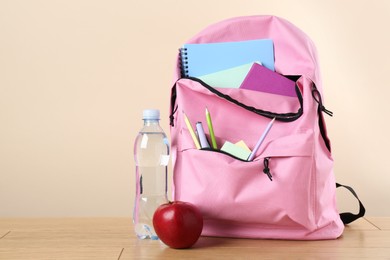 Backpack with different school stationery, bottle of water and apple on wooden table against beige background, space for text