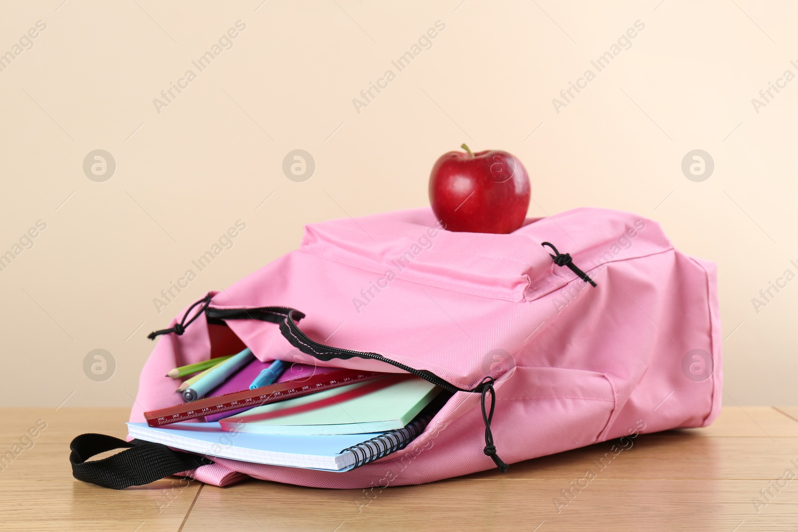 Photo of Backpack with different school stationery and apple on wooden table against beige background