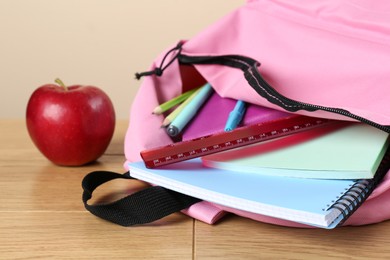 Photo of Backpack with different school stationery and apple on wooden table against beige background, closeup