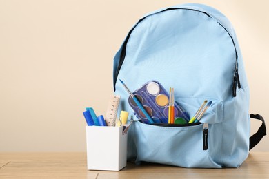 Backpack with different school stationery on wooden table against beige background, space for text