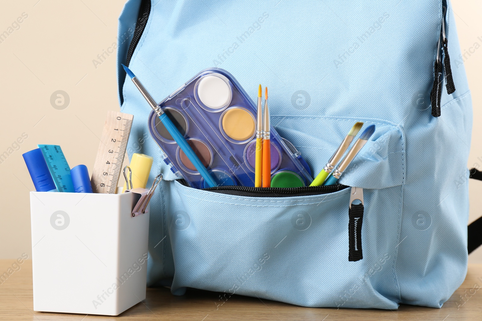 Photo of Backpack with different school stationery on wooden table against beige background, closeup