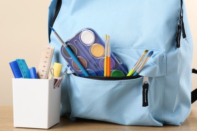 Backpack with different school stationery on wooden table against beige background, closeup