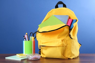 Backpack with different school stationery on wooden table against blue background