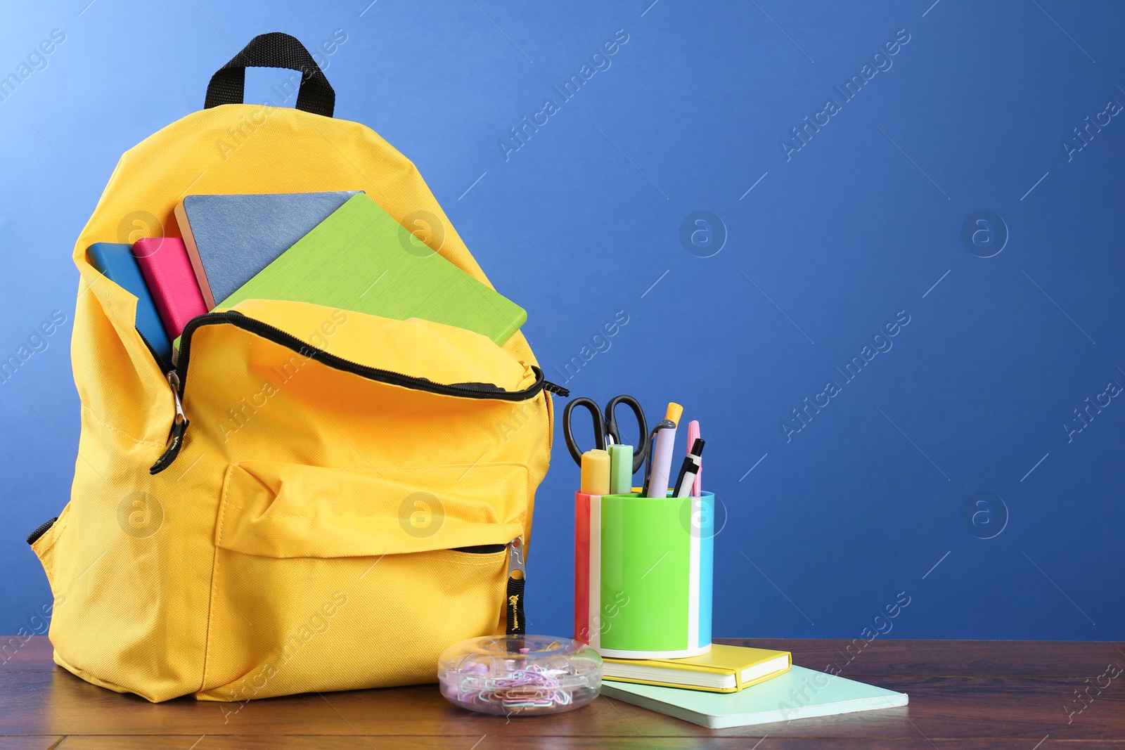 Photo of Backpack with different school stationery on wooden table against blue background, space for text