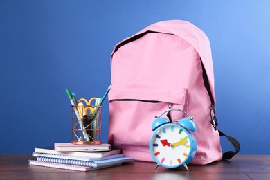 Photo of Backpack with different school stationery and alarm clock on wooden table against blue background