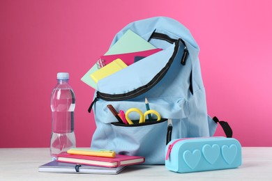 Backpack with different school stationery and bottle of water on light table against pink background