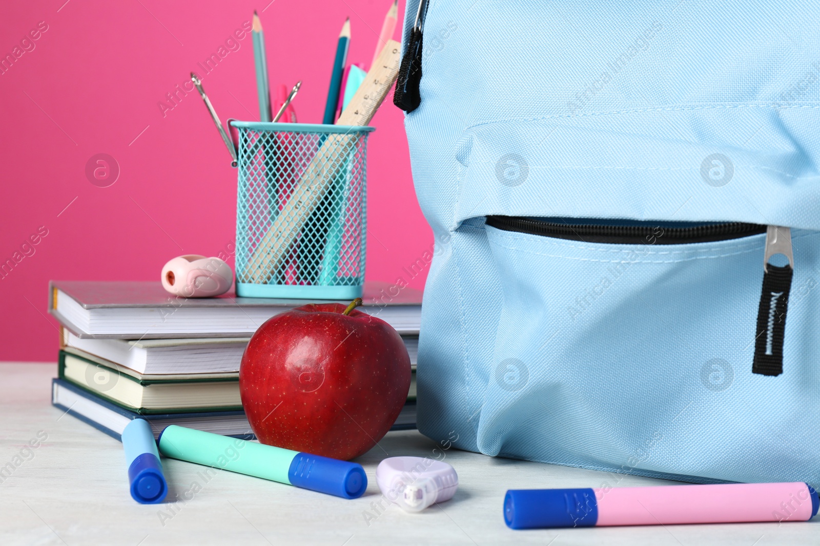 Photo of Backpack with different school stationery and apple on light table against pink background, closeup