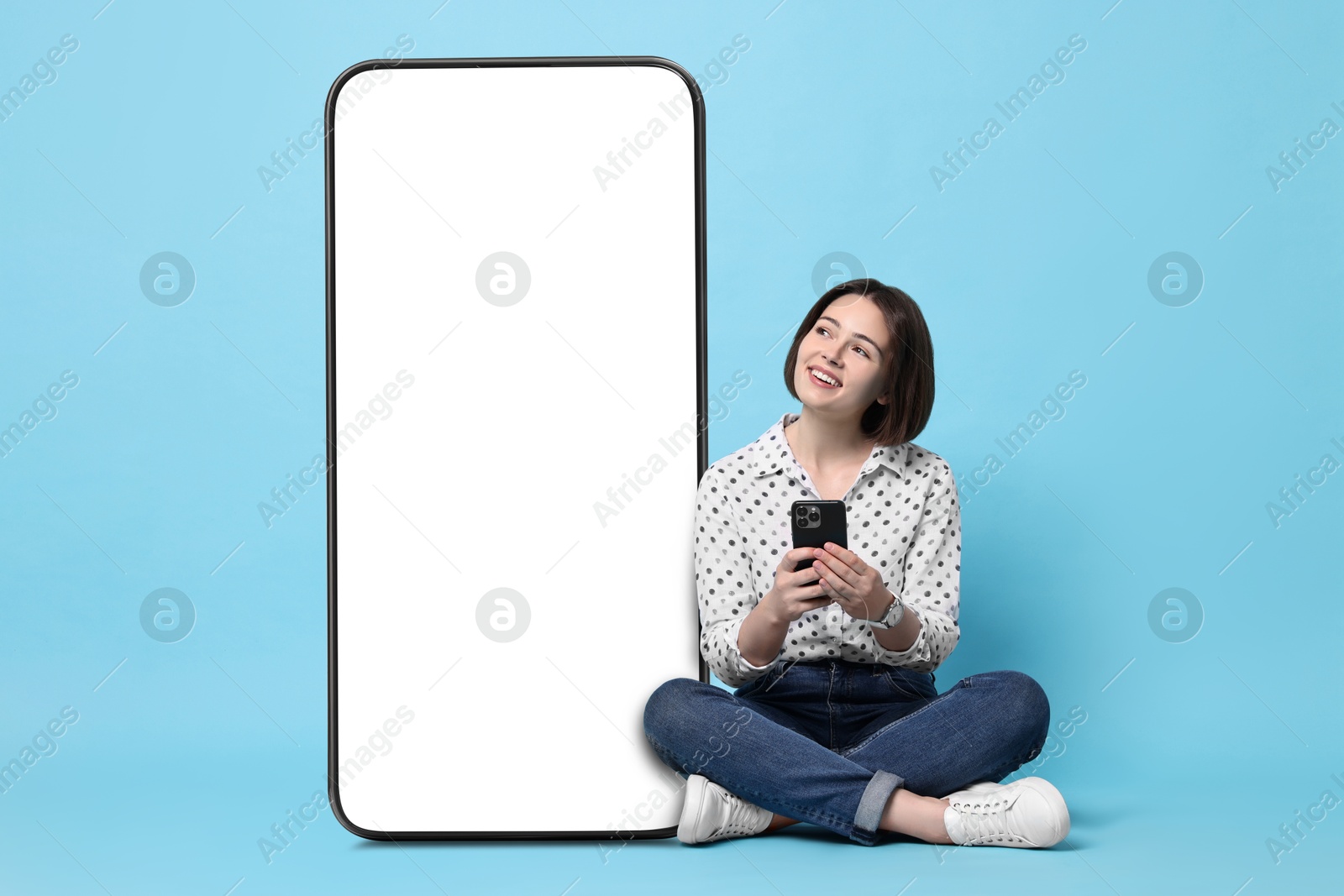 Image of Beautiful young woman with smartphone sitting near big mobile phone with blank screen on light blue background