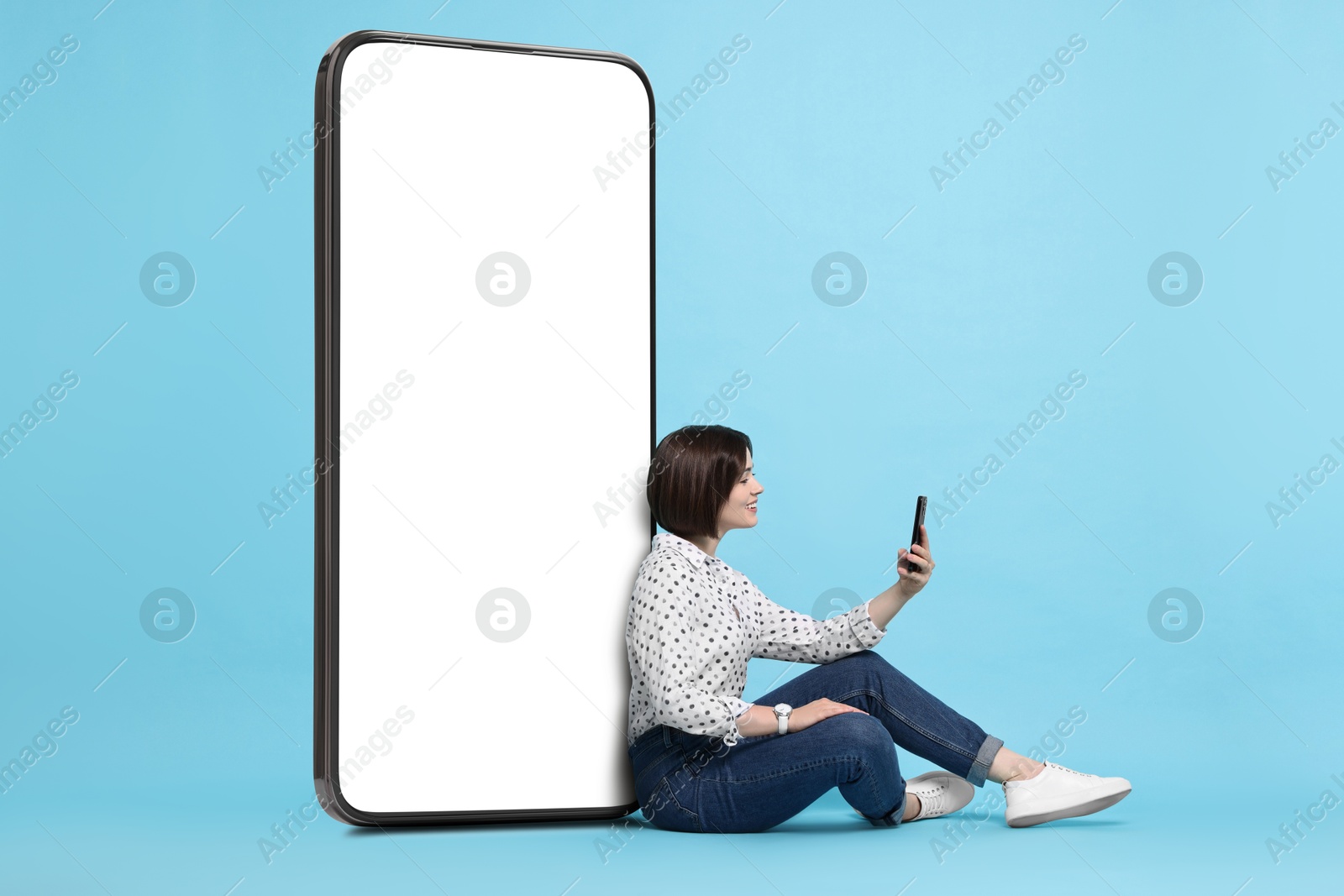 Image of Beautiful young woman with smartphone sitting near big mobile phone with blank screen on light blue background