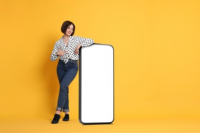 Image of Beautiful young woman pointing at big mobile phone with blank screen on golden background