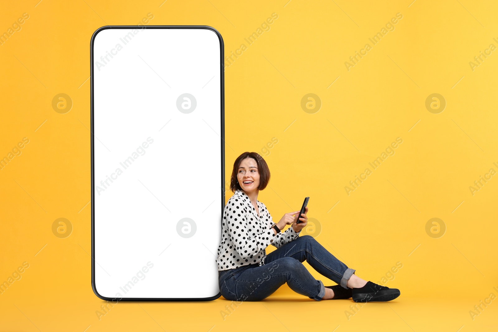 Image of Beautiful young woman with smartphone sitting near big mobile phone with blank screen on golden background