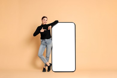 Image of Beautiful young woman pointing at big mobile phone with blank screen on dark beige background