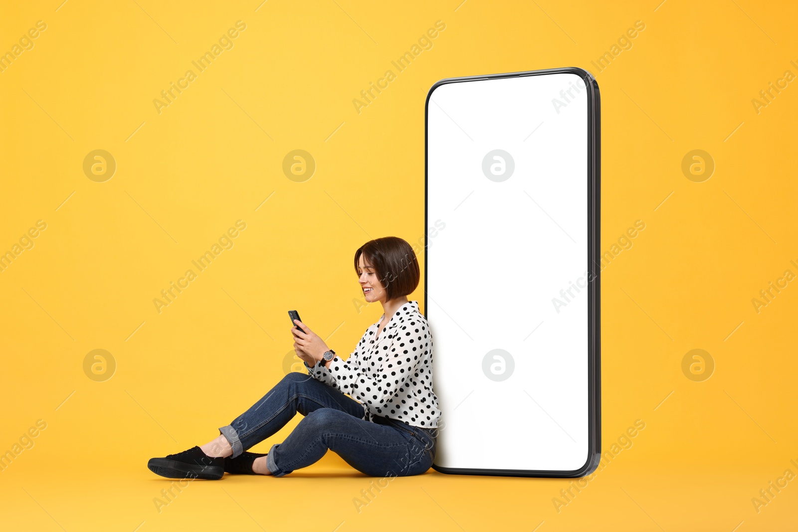 Image of Beautiful young woman with smartphone sitting near big mobile phone with blank screen on golden background