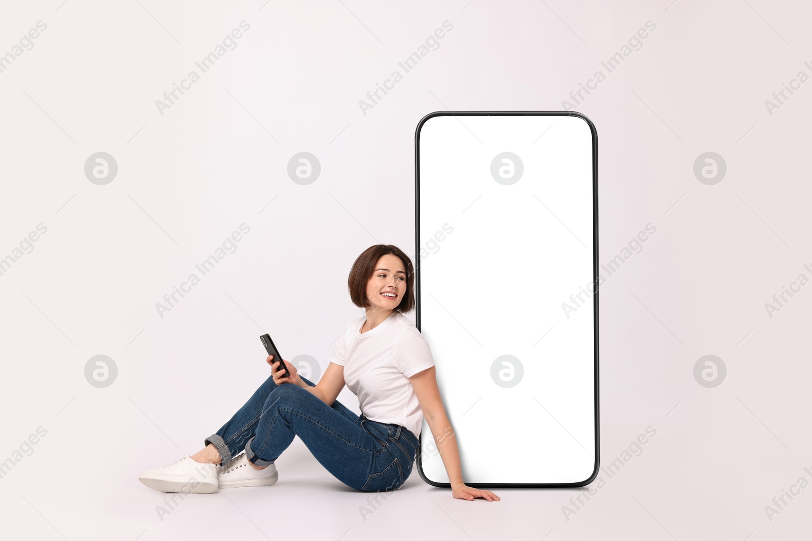 Image of Beautiful young woman with smartphone sitting near big mobile phone with blank screen on light background