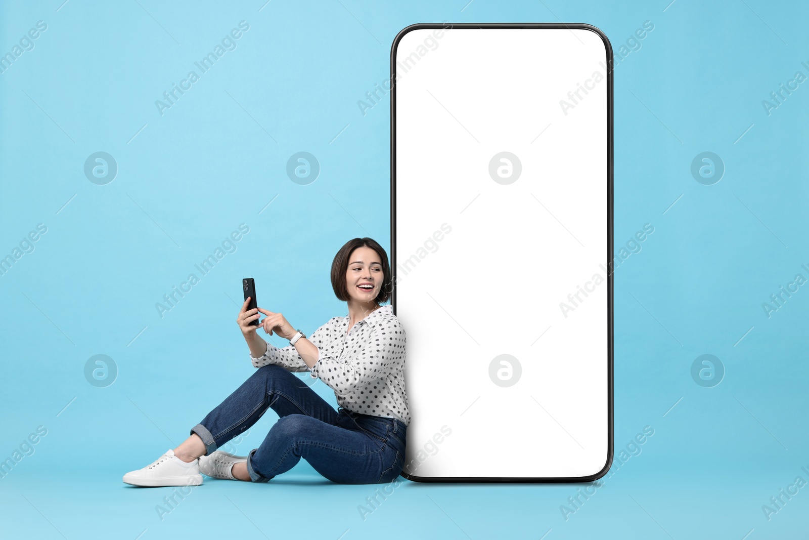 Image of Beautiful young woman with smartphone sitting near big mobile phone with blank screen on light blue background