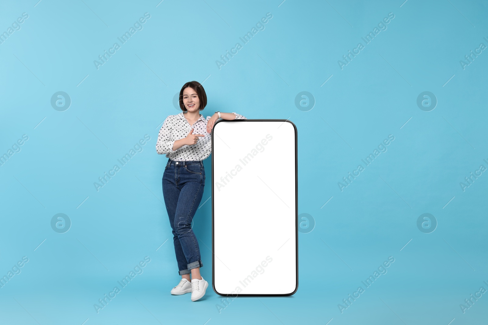 Image of Beautiful young woman pointing at big mobile phone with blank screen on light blue background