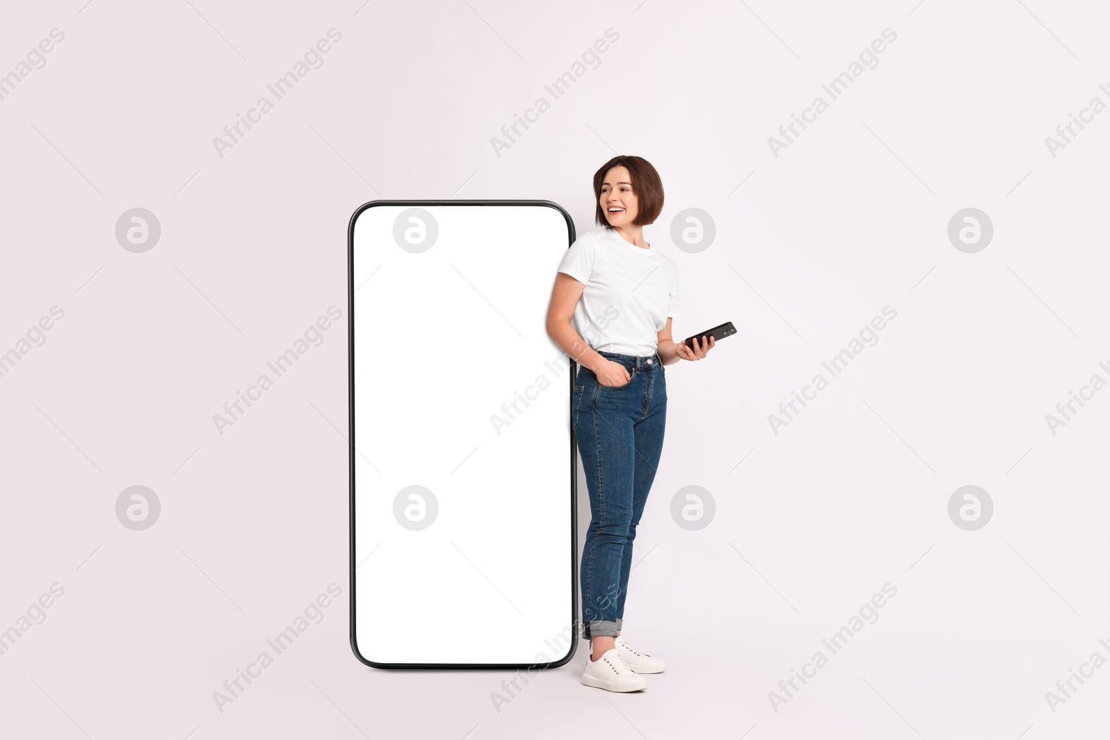 Image of Beautiful young woman with smartphone standing near big mobile phone with blank screen on light background