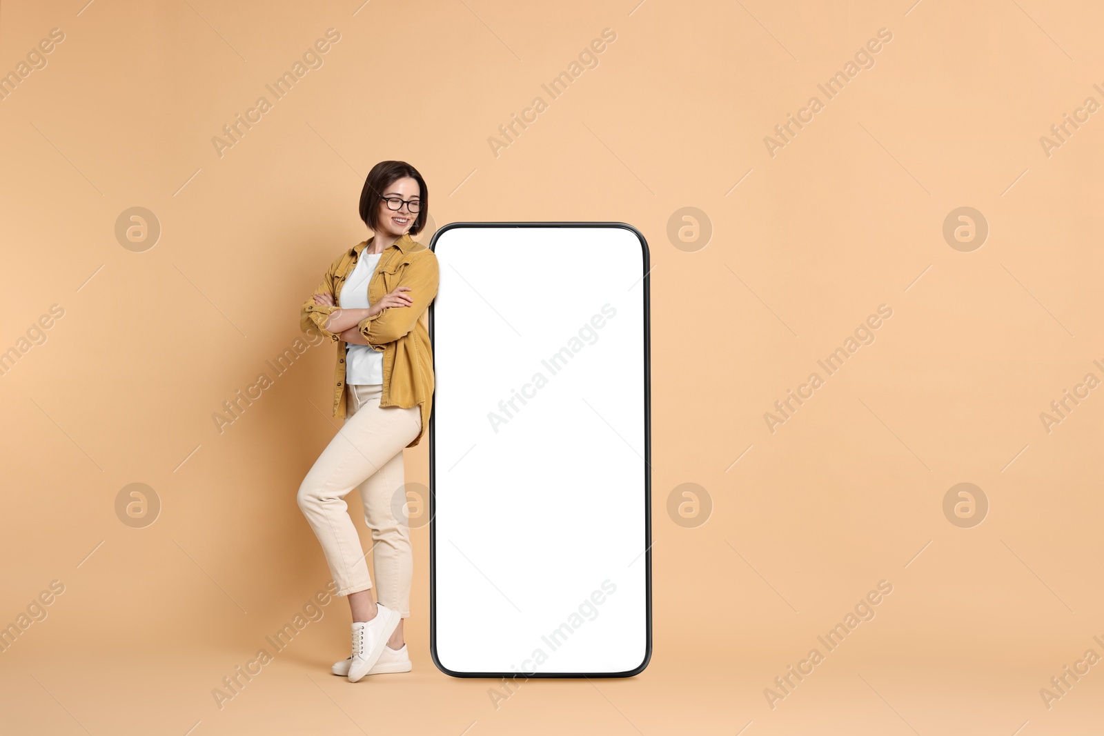 Image of Beautiful young woman standing near big mobile phone with blank screen on dark beige background