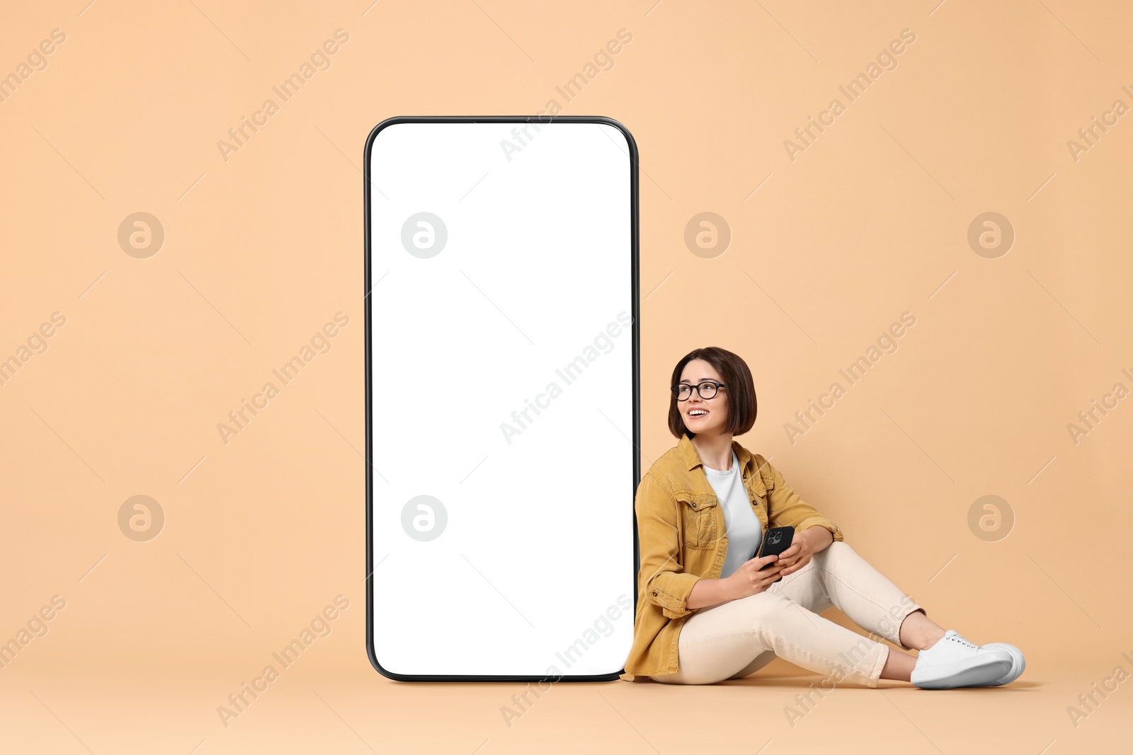 Image of Beautiful young woman with smartphone sitting near big mobile phone with blank screen on dark beige background