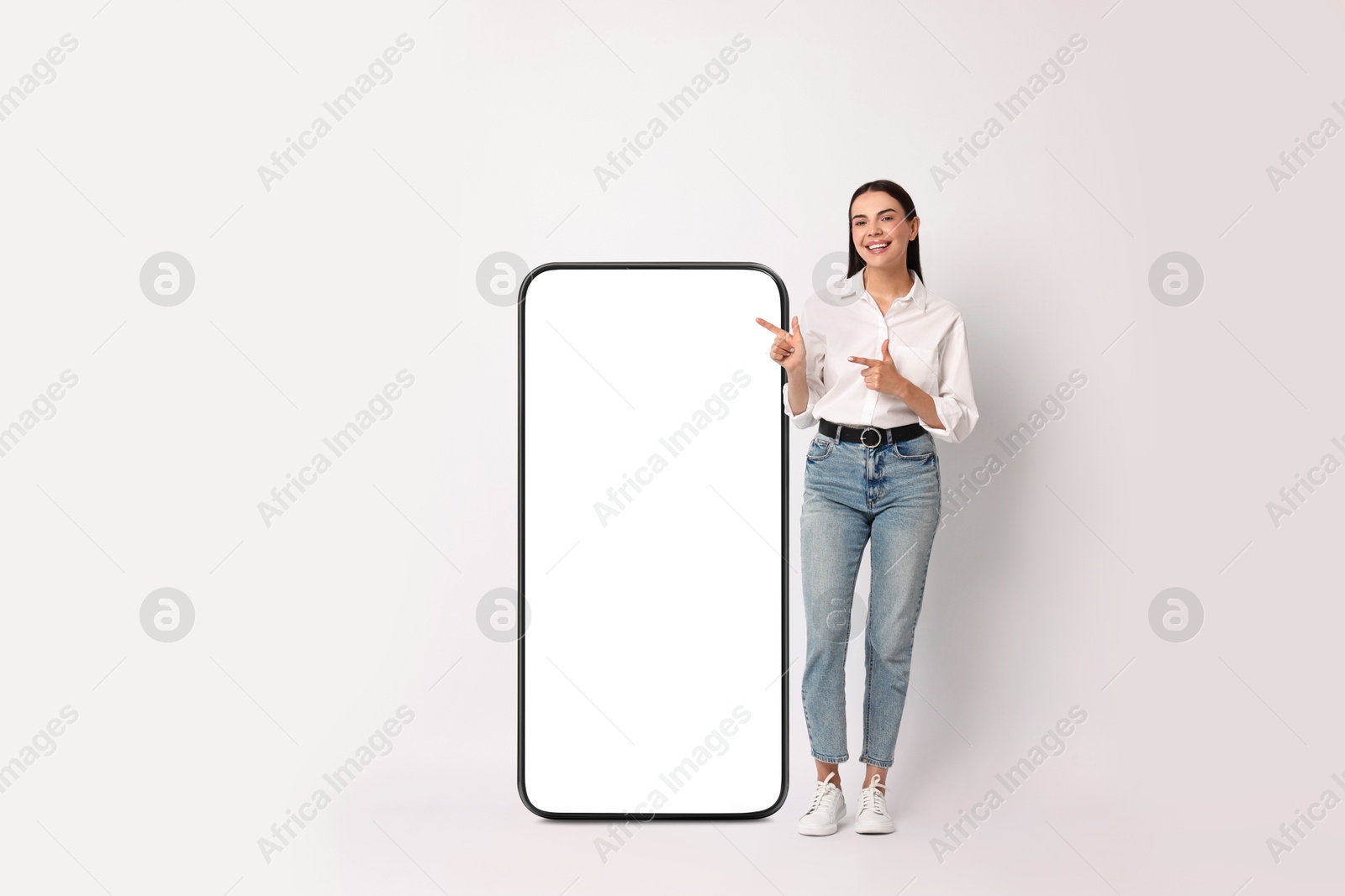 Image of Beautiful young woman pointing at big mobile phone with blank screen on light background