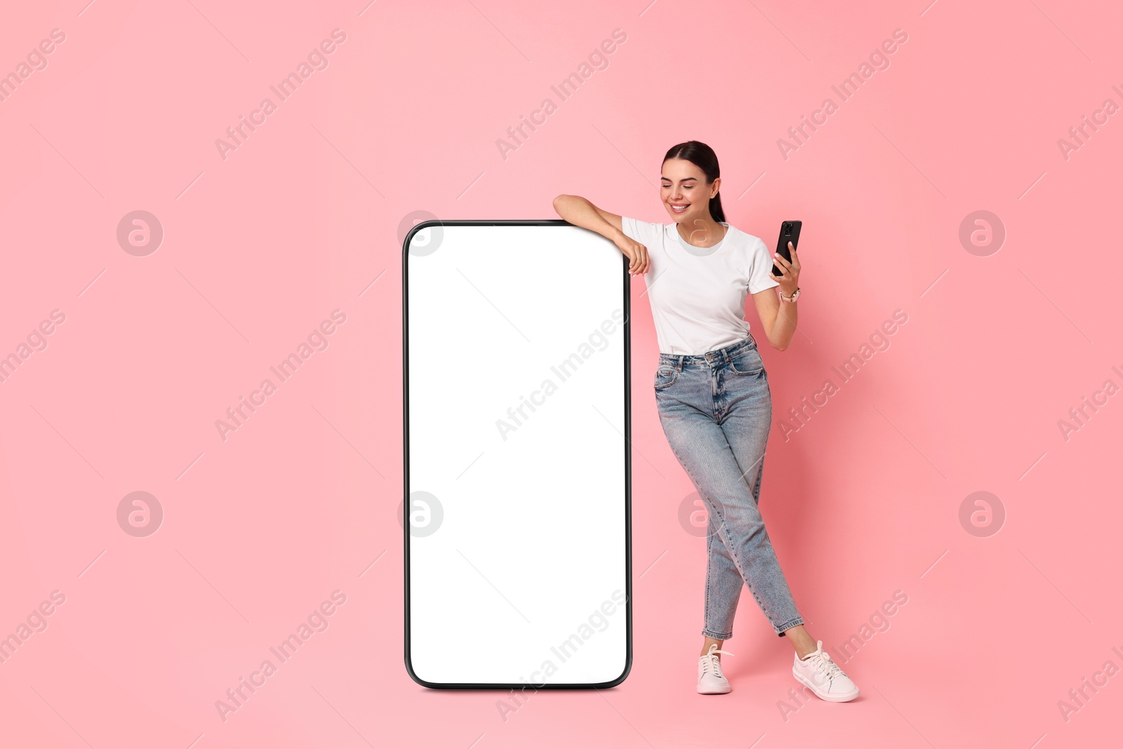 Image of Beautiful young woman with smartphone standing near big mobile phone with blank screen on pink background