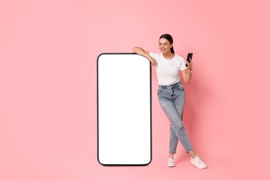 Beautiful young woman with smartphone standing near big mobile phone with blank screen on pink background