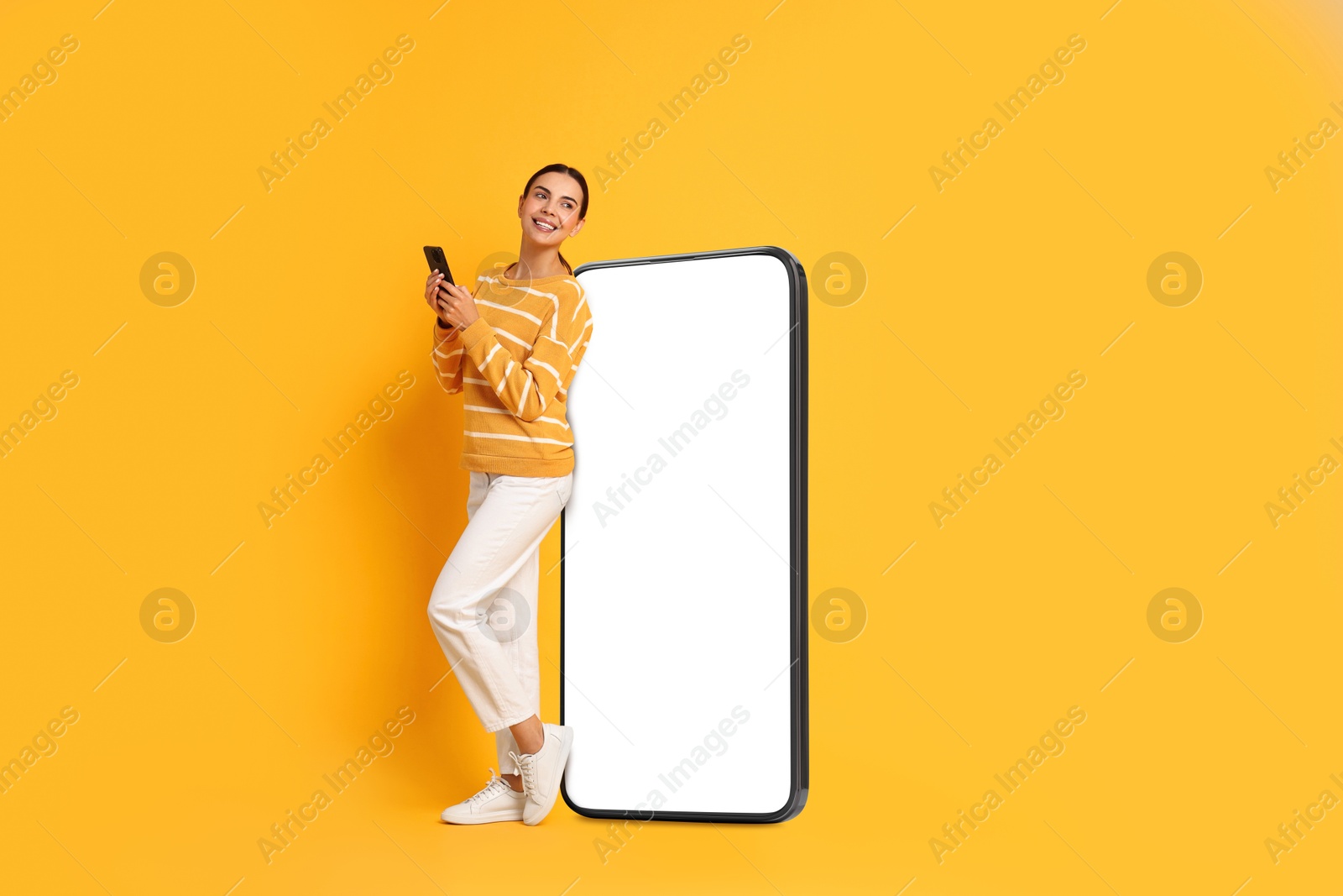 Image of Beautiful young woman with smartphone standing near big mobile phone with blank screen on golden background