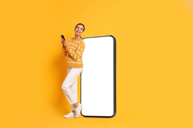 Beautiful young woman with smartphone standing near big mobile phone with blank screen on golden background