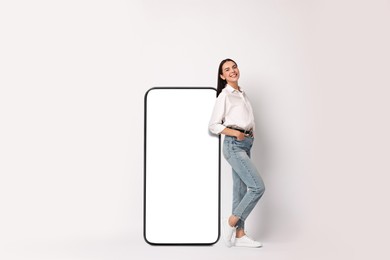 Image of Beautiful young woman standing near big mobile phone with blank screen on light background