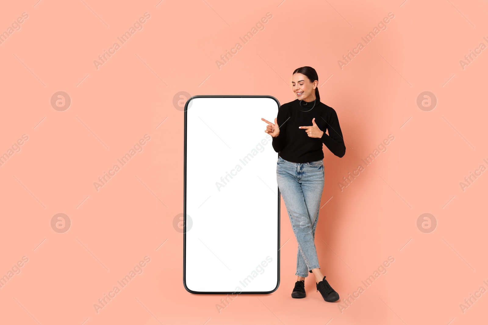 Image of Beautiful young woman pointing at big mobile phone with blank screen on light coral background