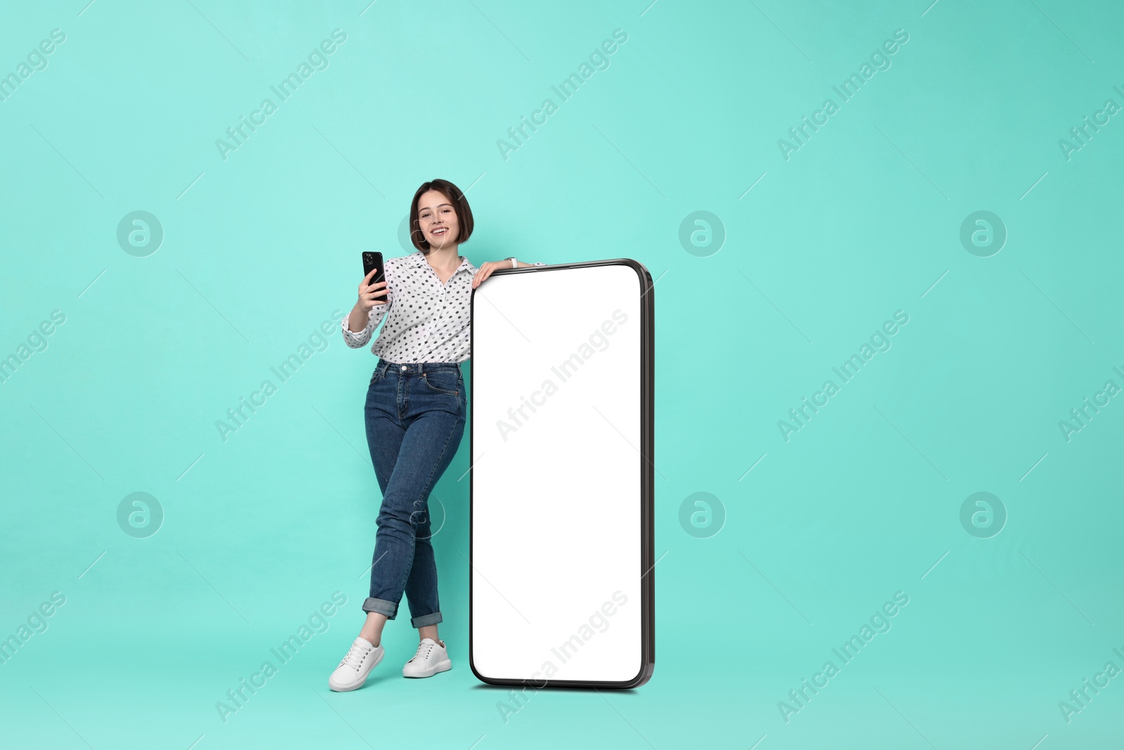 Image of Beautiful young woman with smartphone standing near big mobile phone with blank screen on turquoise background