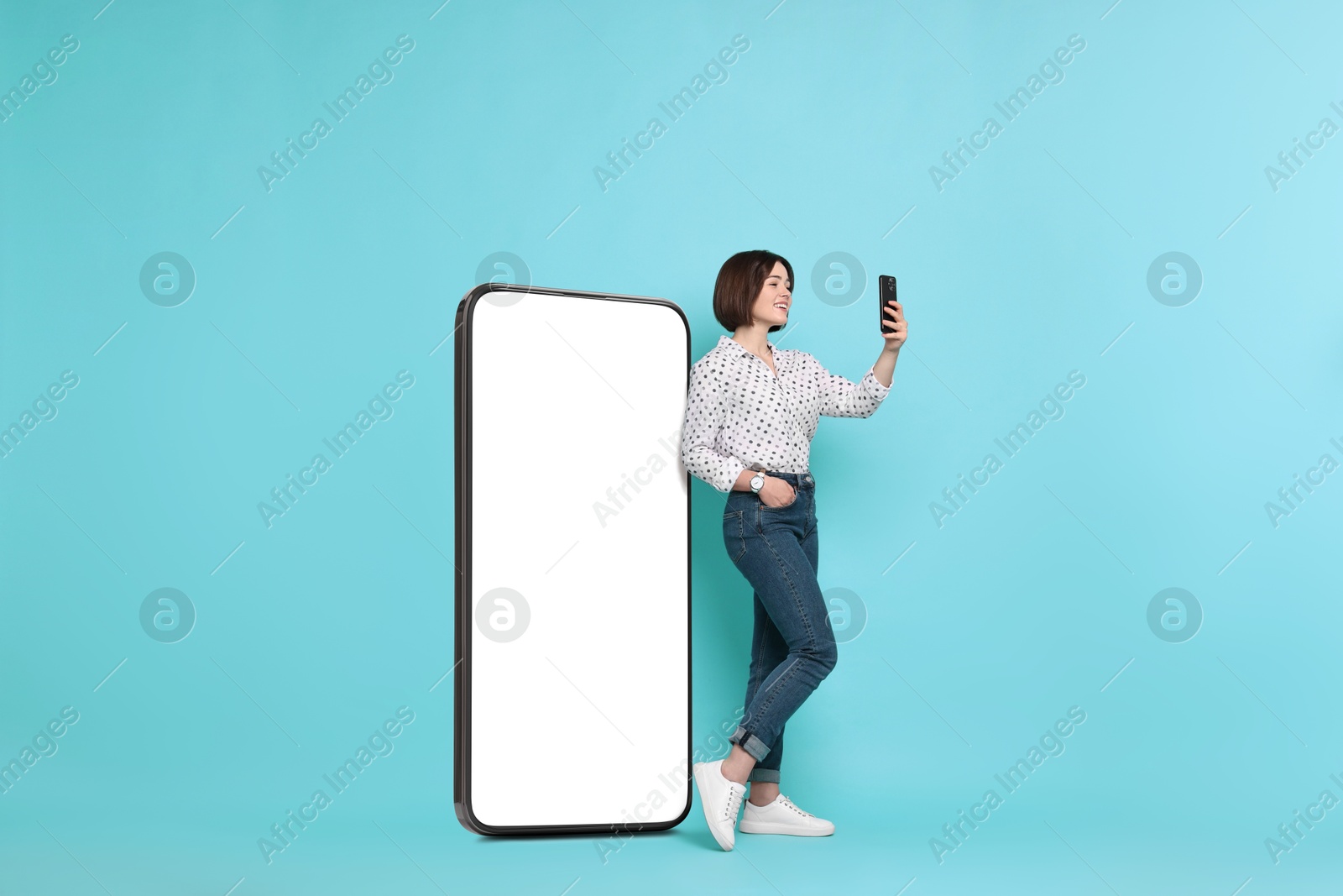 Image of Beautiful young woman with smartphone standing near big mobile phone with blank screen on light blue background