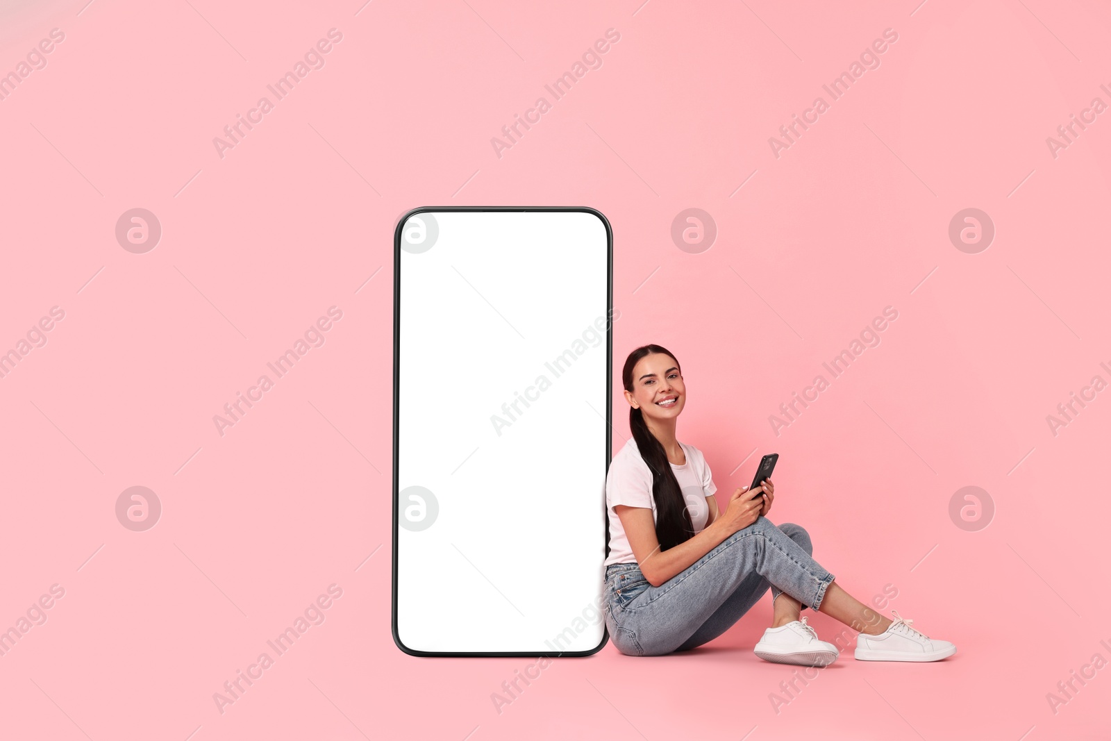 Image of Beautiful young woman with smartphone sitting near big mobile phone with blank screen on pink background