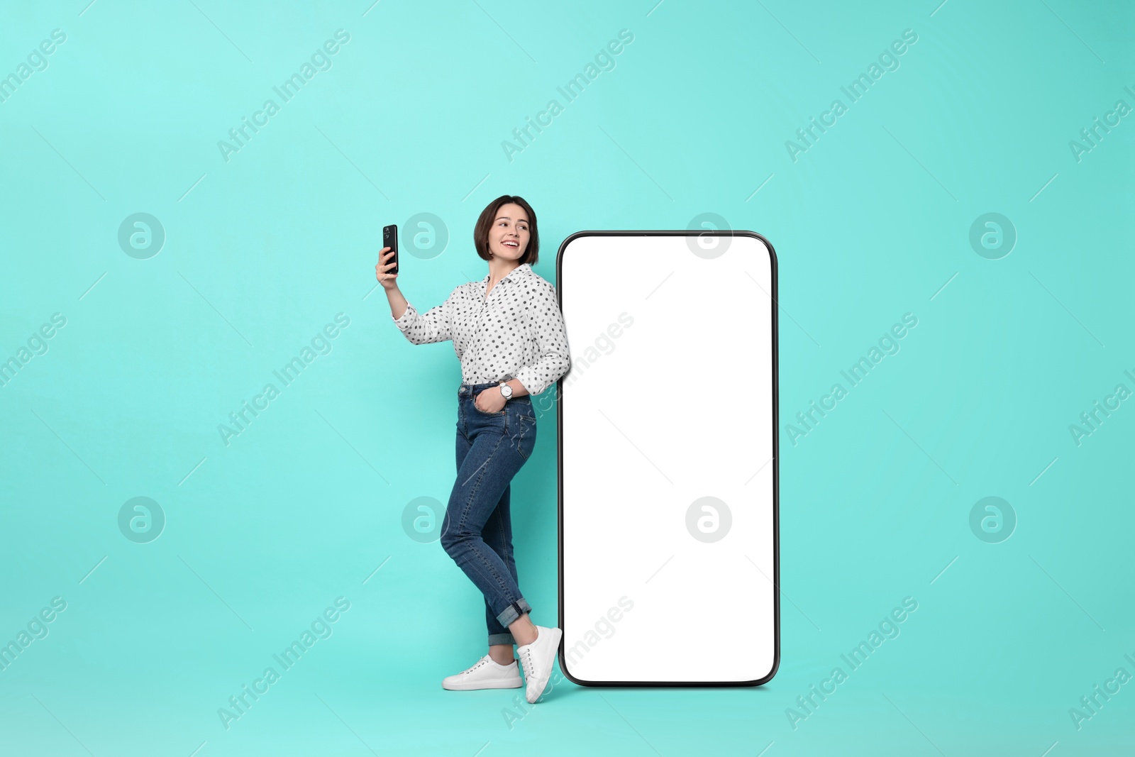 Image of Beautiful young woman with smartphone standing near big mobile phone with blank screen on turquoise background