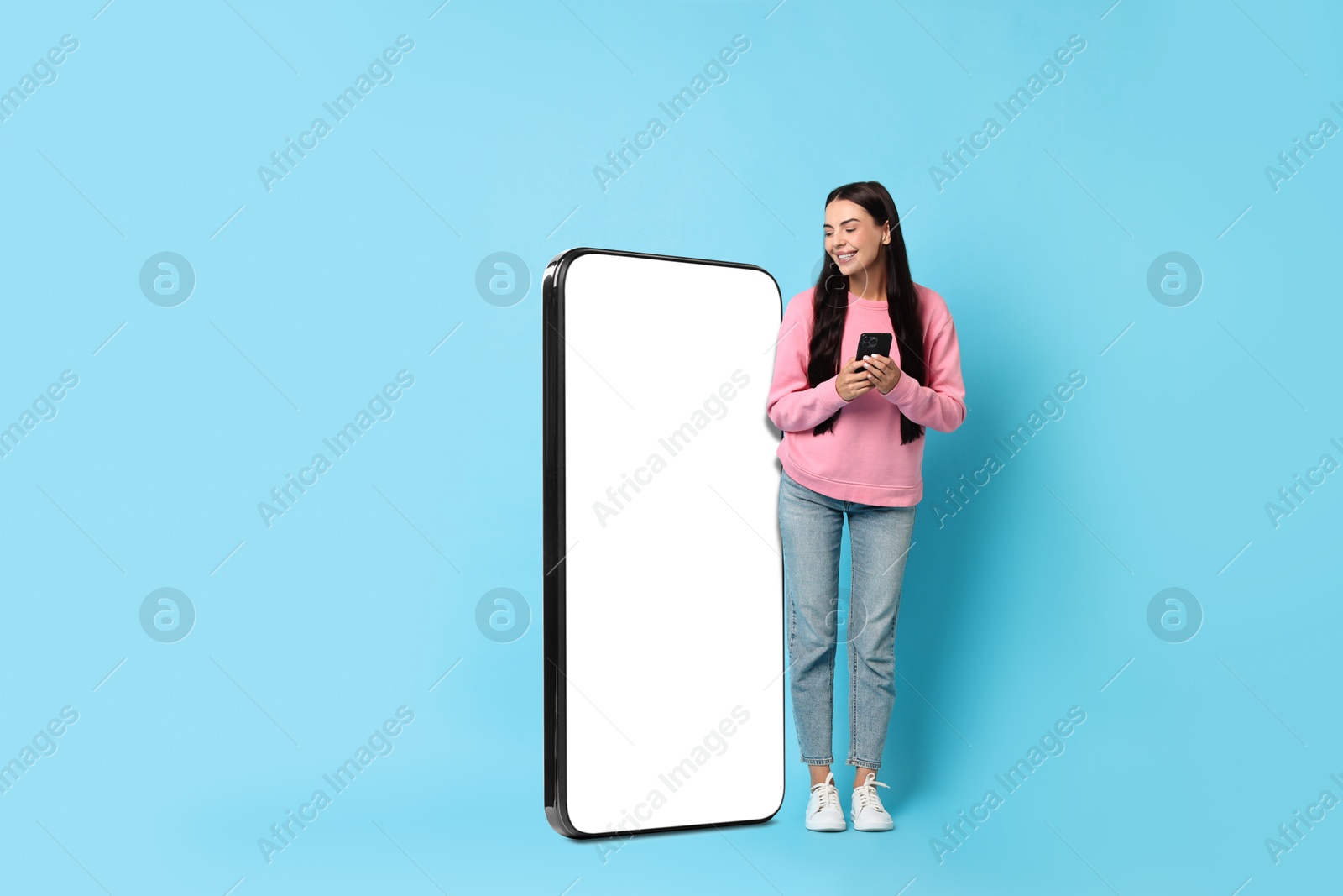 Image of Beautiful young woman with smartphone standing near big mobile phone with blank screen on light blue background