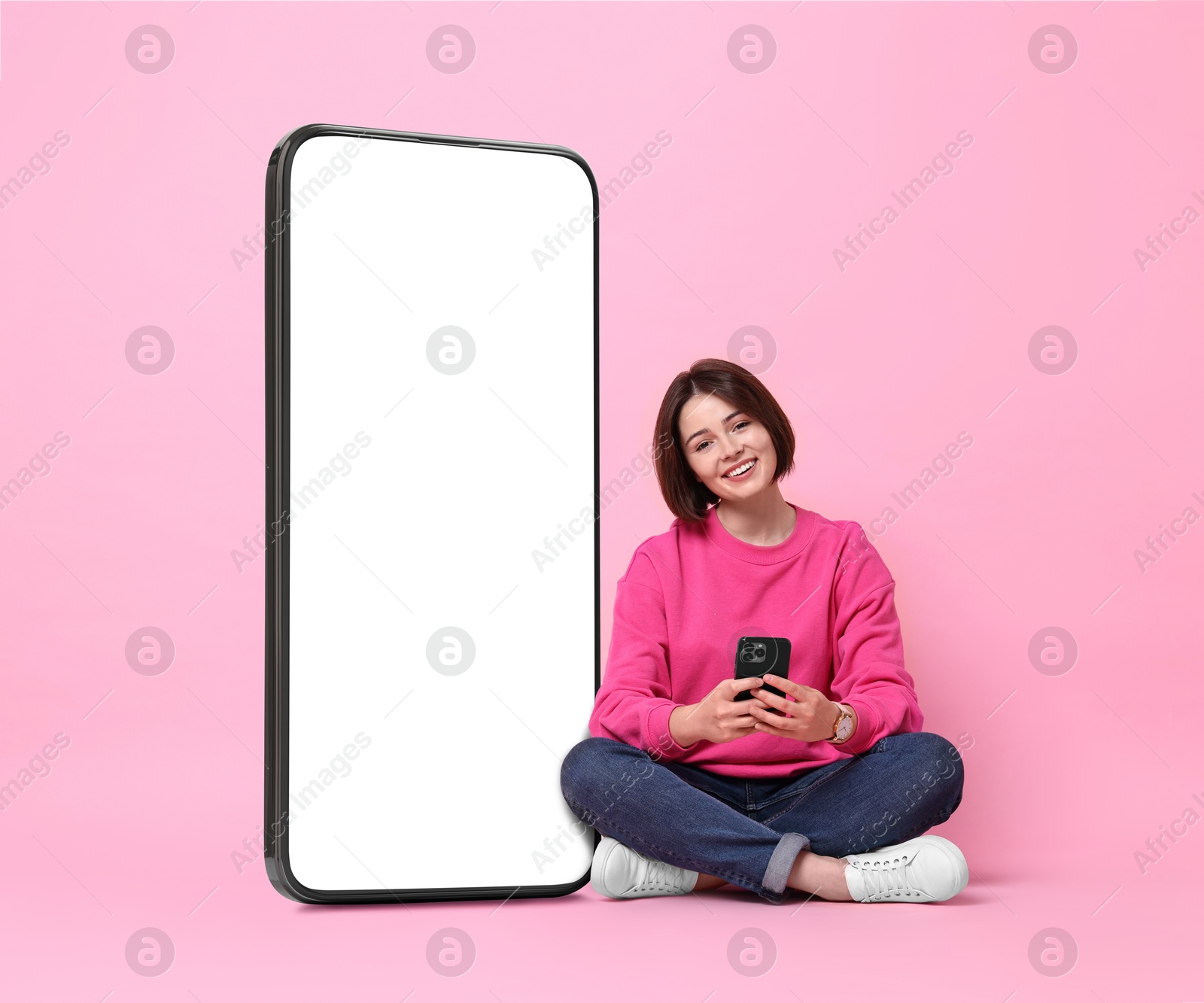 Image of Beautiful young woman with smartphone sitting near big mobile phone with blank screen on pink background