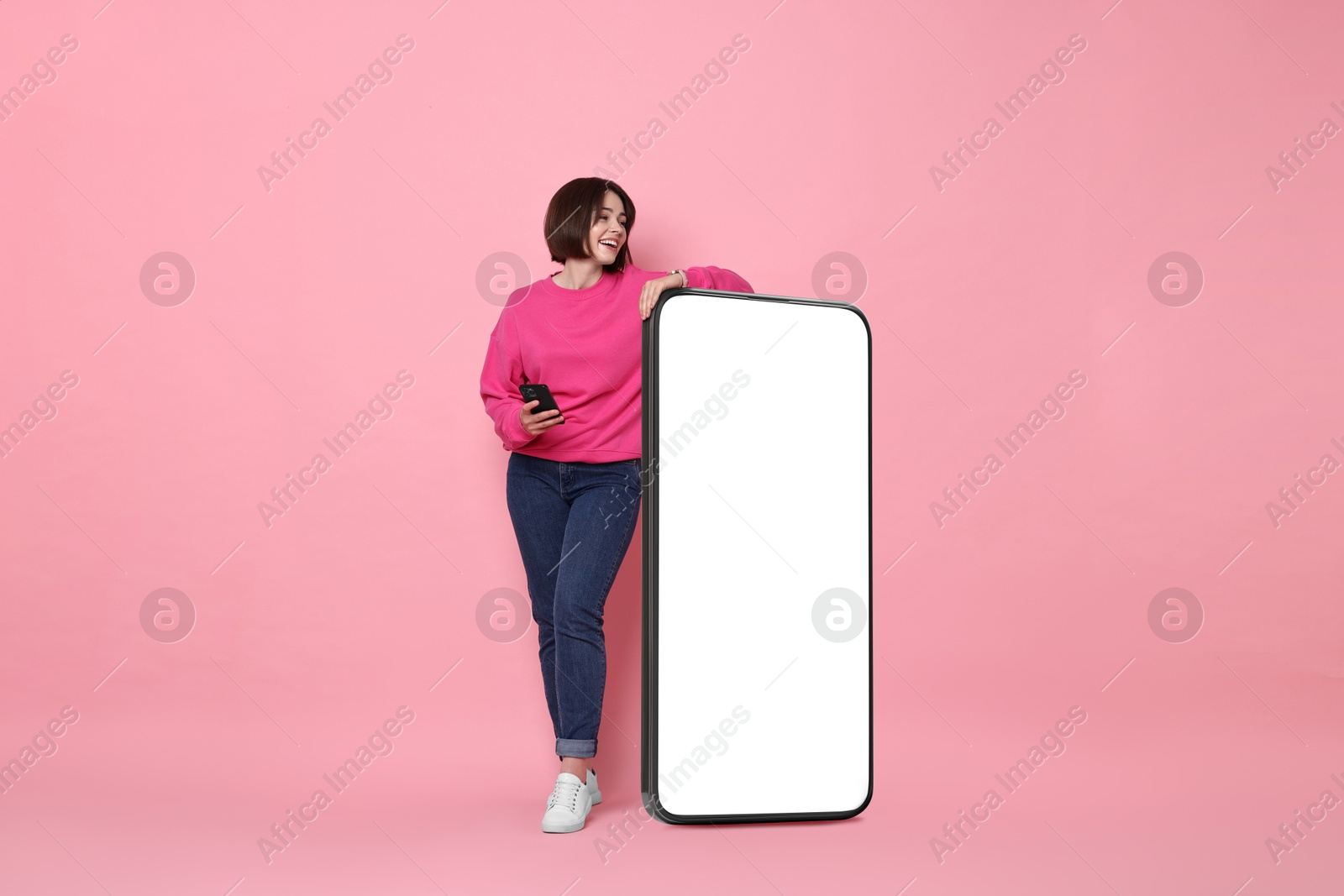 Image of Beautiful young woman standing near big mobile phone with blank screen on pink background