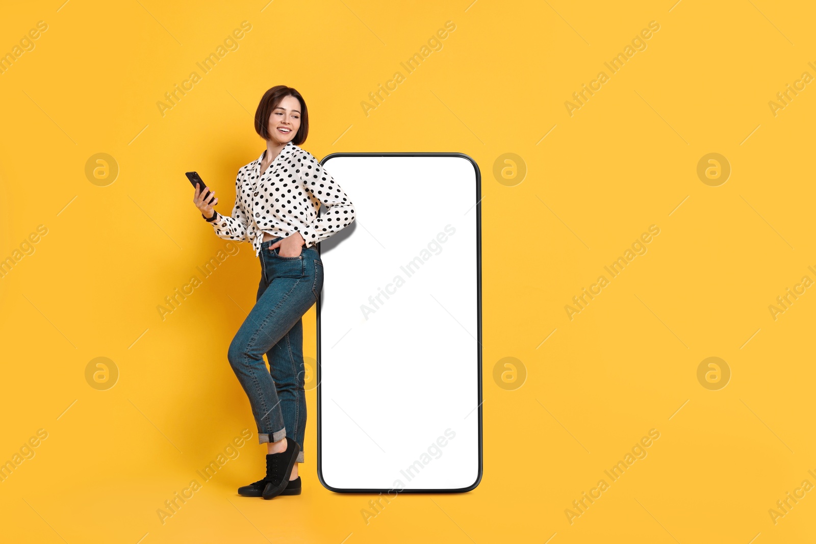 Image of Beautiful young woman with smartphone standing near big mobile phone with blank screen on golden background