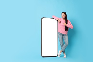 Beautiful young woman pointing at big mobile phone with blank screen on light blue background