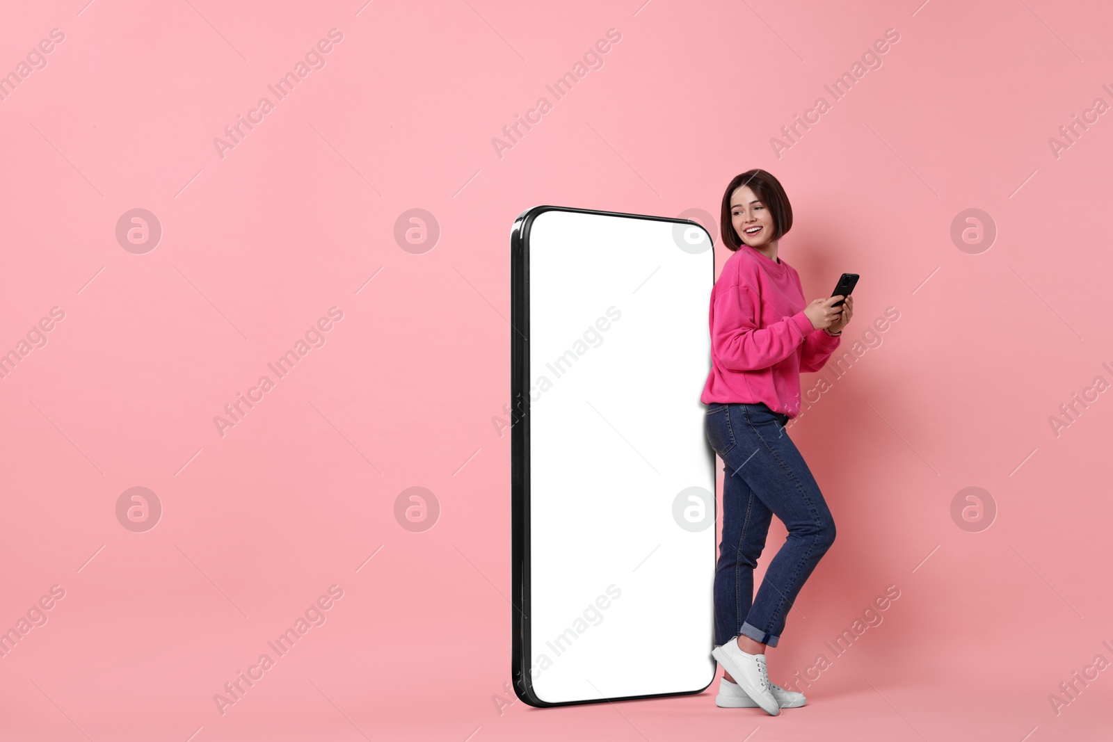 Image of Beautiful young woman with smartphone standing near big mobile phone with blank screen on pink background