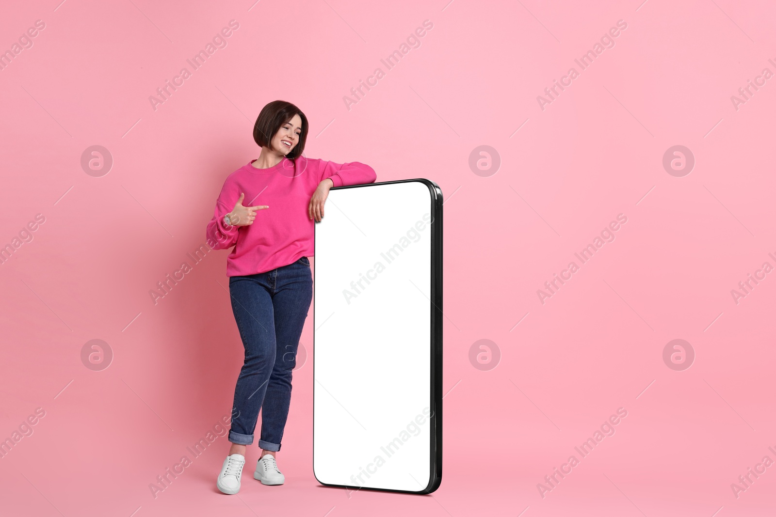 Image of Beautiful young woman pointing at big mobile phone with blank screen on pink background