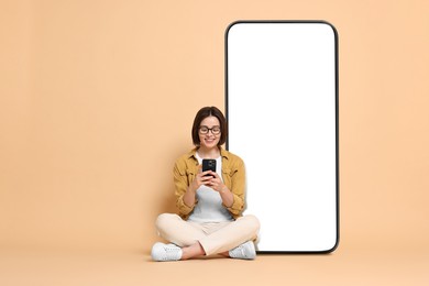 Beautiful young woman with smartphone sitting near big mobile phone with blank screen on dark beige background