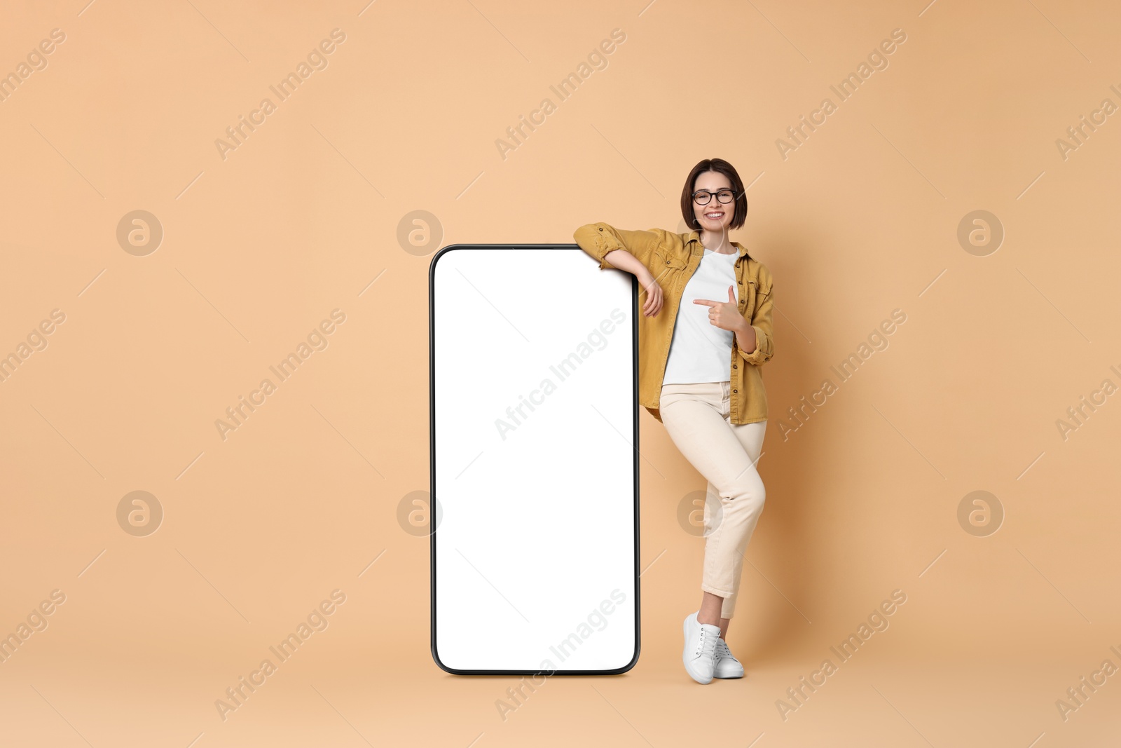 Image of Beautiful young woman pointing at big mobile phone with blank screen on dark beige background