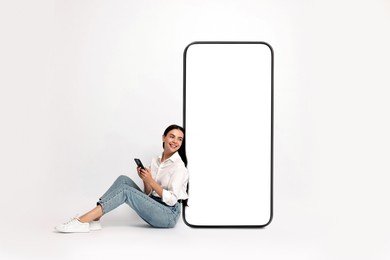 Beautiful young woman with smartphone sitting near big mobile phone with blank screen on light background