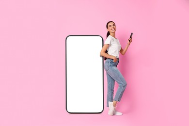 Beautiful young woman with smartphone standing near big mobile phone with blank screen on pink background