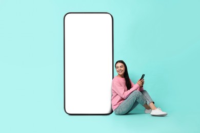 Image of Beautiful young woman with smartphone sitting near big mobile phone with blank screen on turquoise background