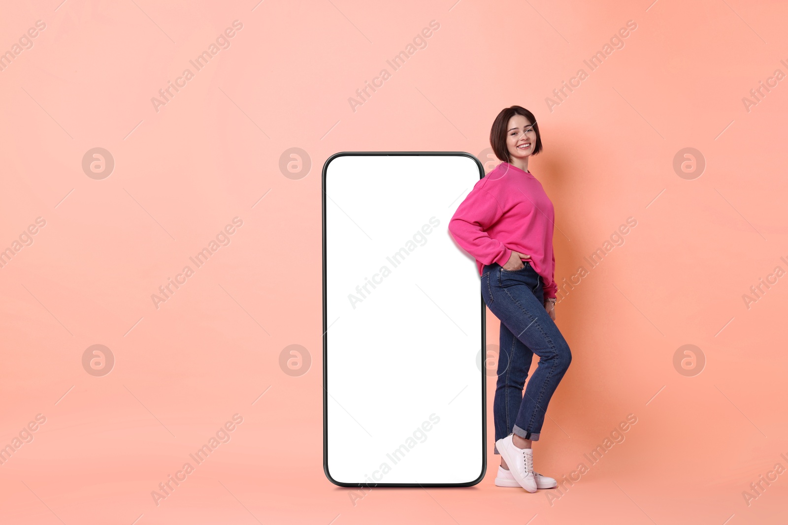 Image of Beautiful young woman standing near big mobile phone with blank screen on light coral background