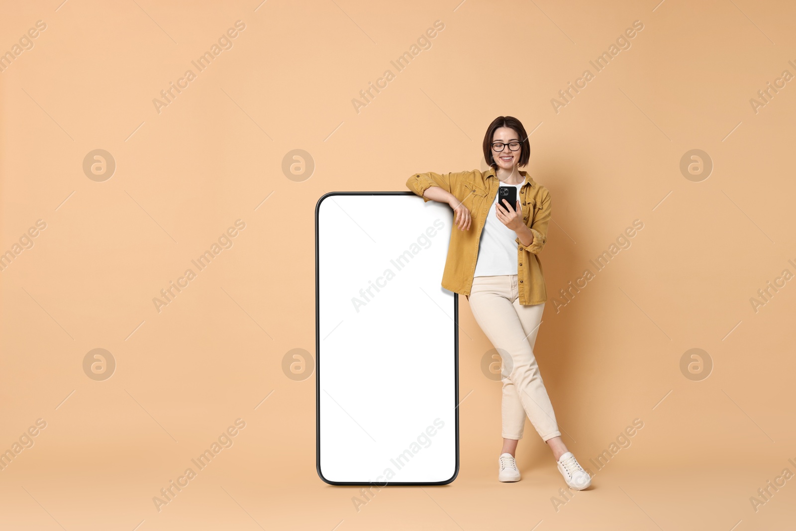 Image of Beautiful young woman with smartphone standing near big mobile phone with blank screen on dark beige background