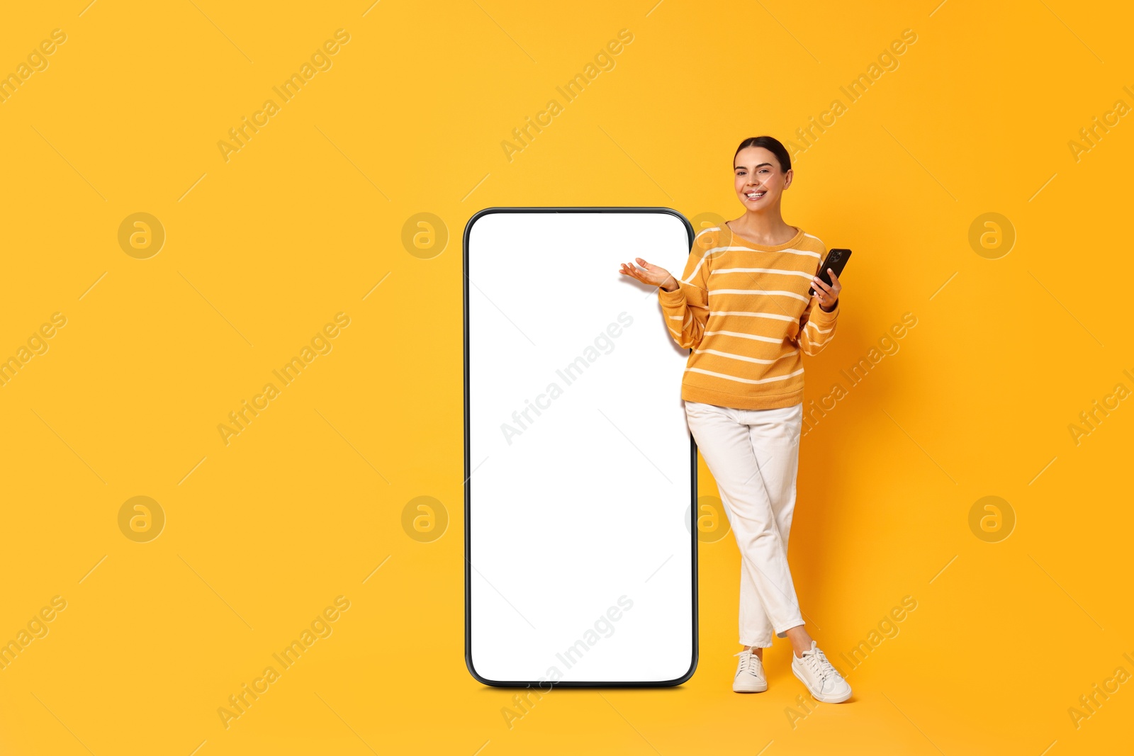 Image of Beautiful young woman with smartphone standing near big mobile phone with blank screen on golden background